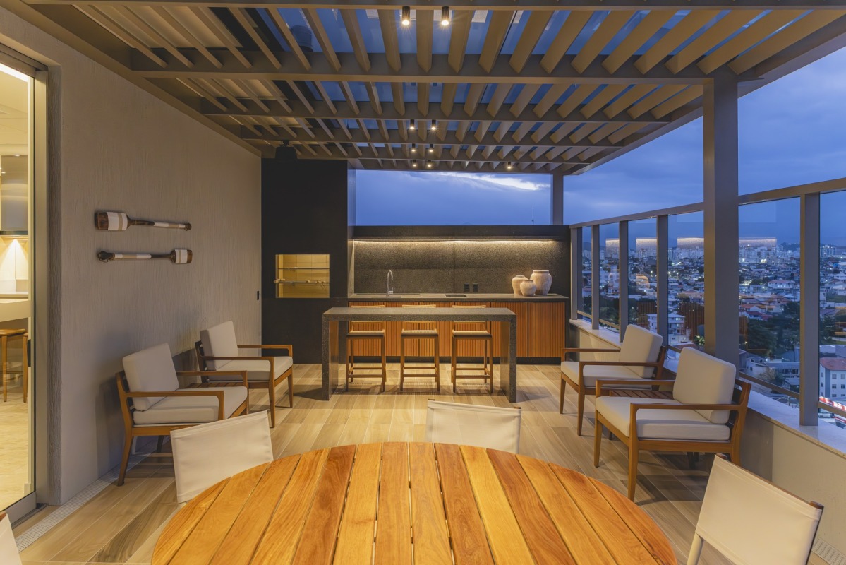 Varanda gourmet moderna em uma cobertura ao anoitecer, com teto de pergolado de madeira, bancada com banquetas, poltronas confortáveis e vista para a cidade.