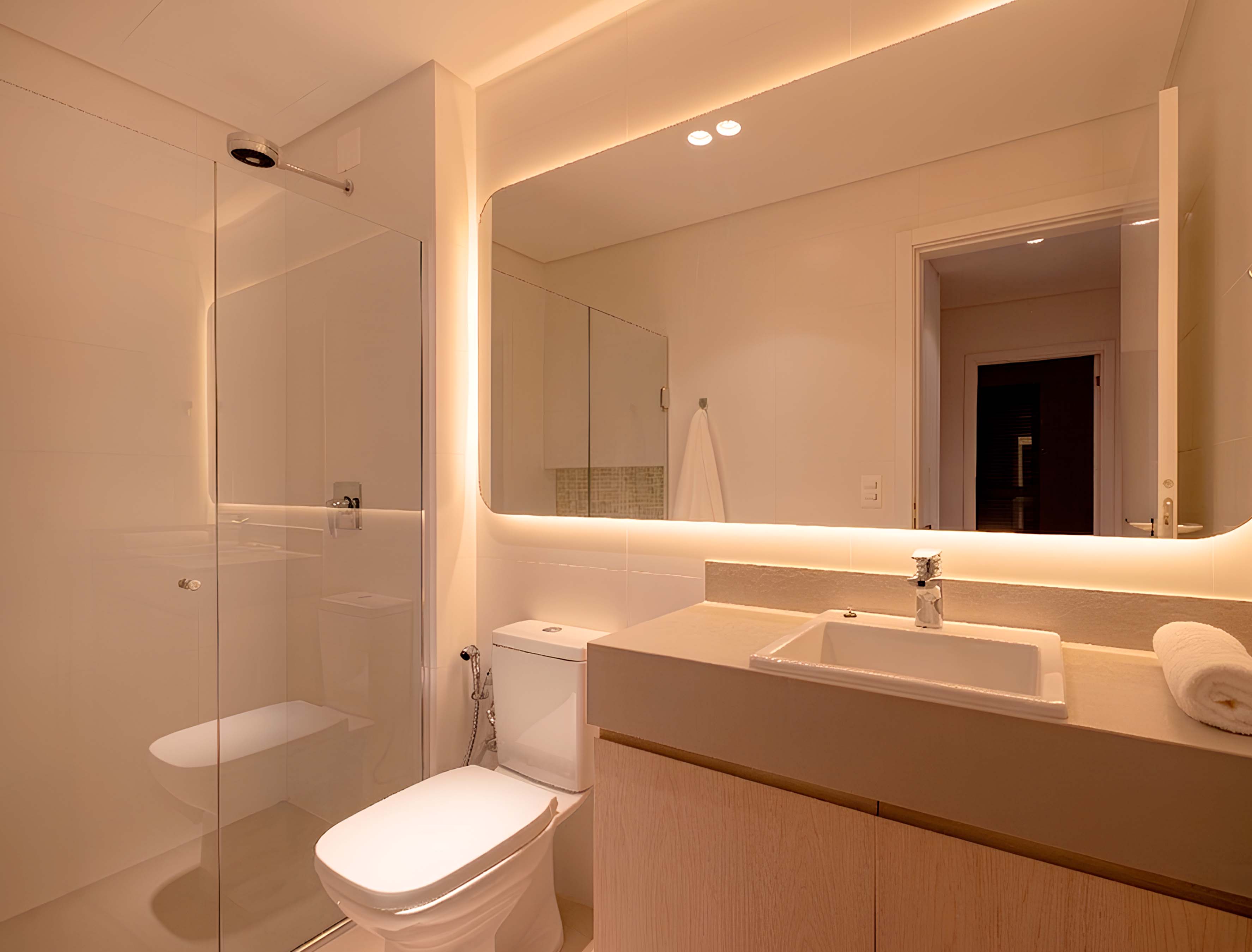 banheiro-planejado-com-box-spa-particular-em-casa.jpg Banheiro moderno com bancada bege, gabinete em madeira clara e espelho com iluminação LED. À esquerda, box de vidro com chuveiro de teto, criando o conceito de um SPA particular em casa.