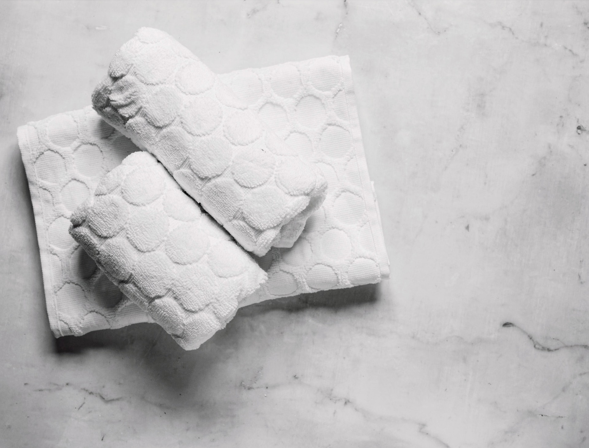 toalhas-algodao-detalhe-spa-particular-casa.jpg Duas toalhas de rosto brancas de algodão, com textura de círculos em relevo, dobradas e empilhadas sobre uma superfície de mármore branco polido com veios cinzas, representando o luxo de um SPA particular em casa.