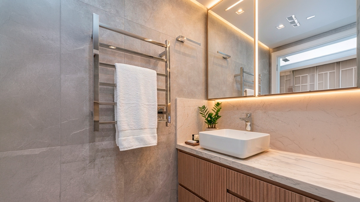 banheiro-luxo-toalheiro-termico-spa-casa.jpg Banheiro de alto padrão com revestimento cinza texturizado e bancada de mármore bege. Em destaque, um toalheiro térmico elétrico em aço cromado com uma toalha branca, ao lado de um espelho com iluminação LED embutida, criando uma atmosfera de SPA particular em casa.