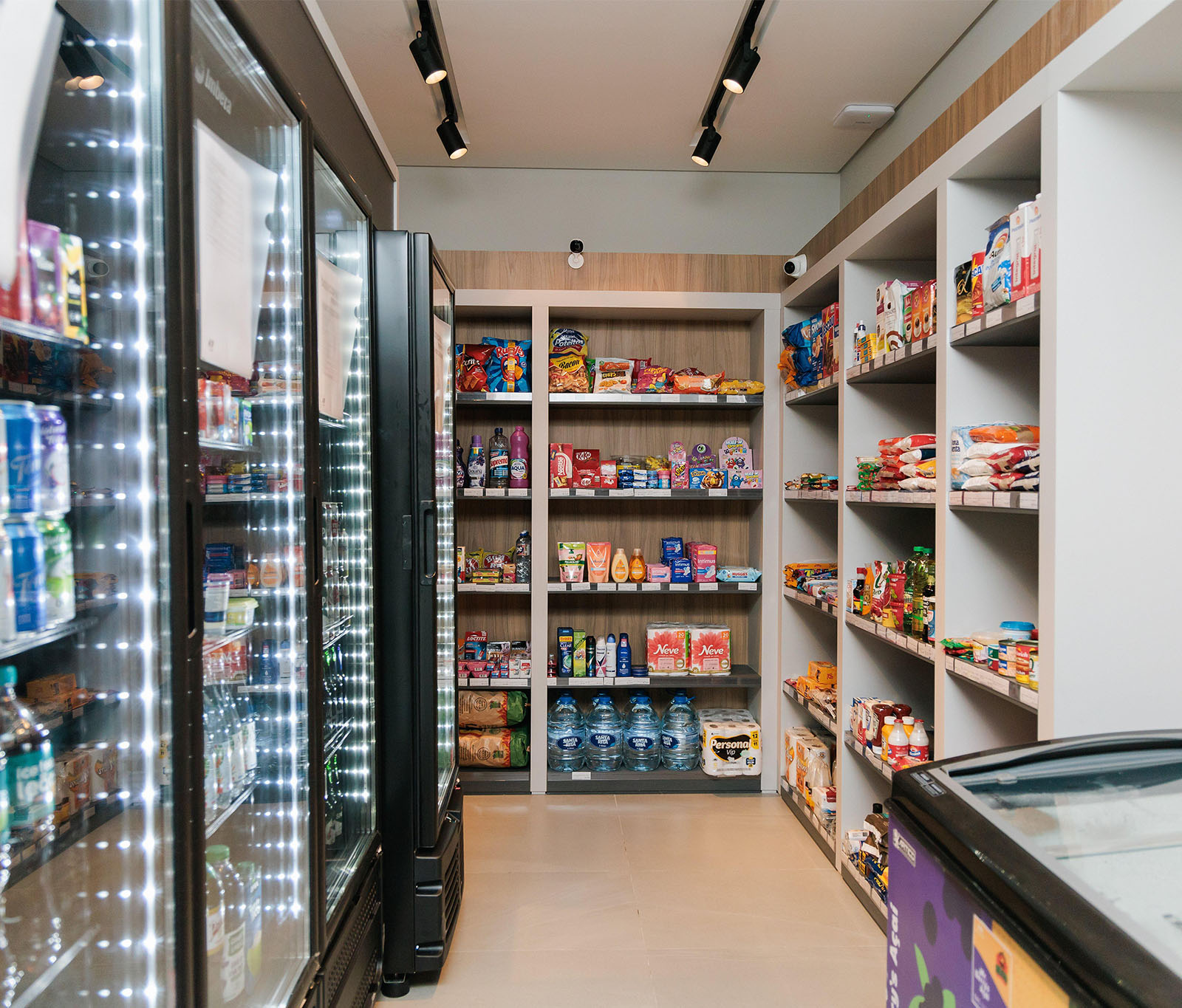 servicos-no-condominio-transformam-sua-rotina.jpg A foto mostra o interior de uma pequena loja de conveniência autônoma. O corredor estreito é ladeado por freezers comerciais altos e prateleiras de madeira. À esquerda, grandes freezers com portas de vidro exibem bebidas frias e alimentos congelados, iluminados por luzes internas de LED. No centro e à direita, prateleiras de madeira e brancas estão cheias de uma variedade de itens, incluindo salgadinhos ensacados, lanches embalados, itens de mercearia e produtos de higiene pessoal. A loja está bem iluminada com iluminação de trilho moderna no teto. O chão é de azulejo bege claro e a loja está limpa e organizada. Serviços de condomínio transformam sua rotina.