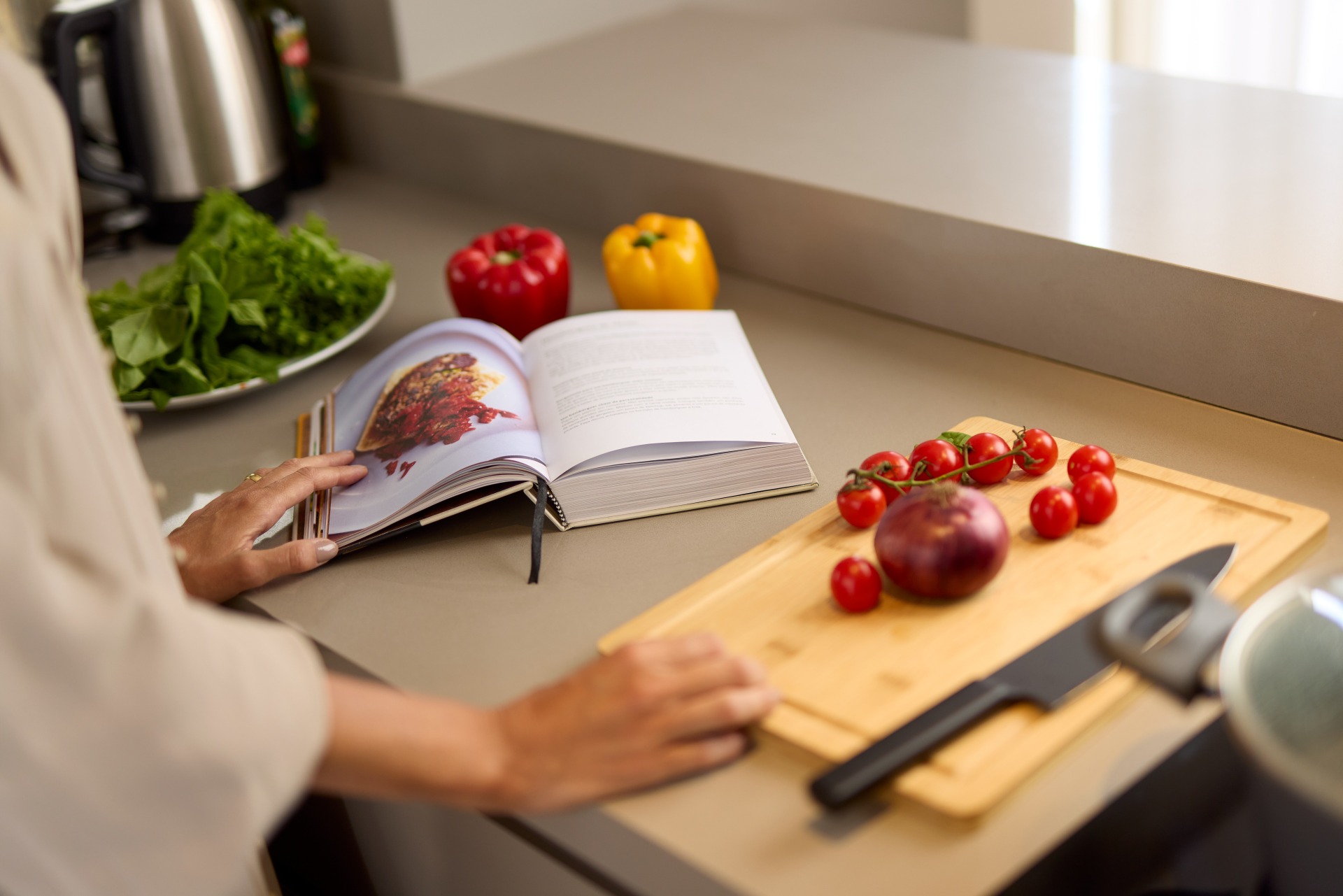 Cozinha semi-profissional em apartamento: Otimizando o espaço para preparo e consulta de receitas Uma vista em close-up de uma pessoa com uma camisa de cor clara de pé em uma bancada de cozinha de cor cinza, lendo um livro de receitas aberto. Suas mãos estão apoiadas na bancada. Ao lado dela, à direita, há uma tábua de corte de madeira com uma faca de chef preta, uma cebola roxa e tomates cereja. Ao fundo, à esquerda, há um prato com verduras frescas, pimentões vermelhos e amarelos inteiros e uma chaleira elétrica. A iluminação é suave e natural.