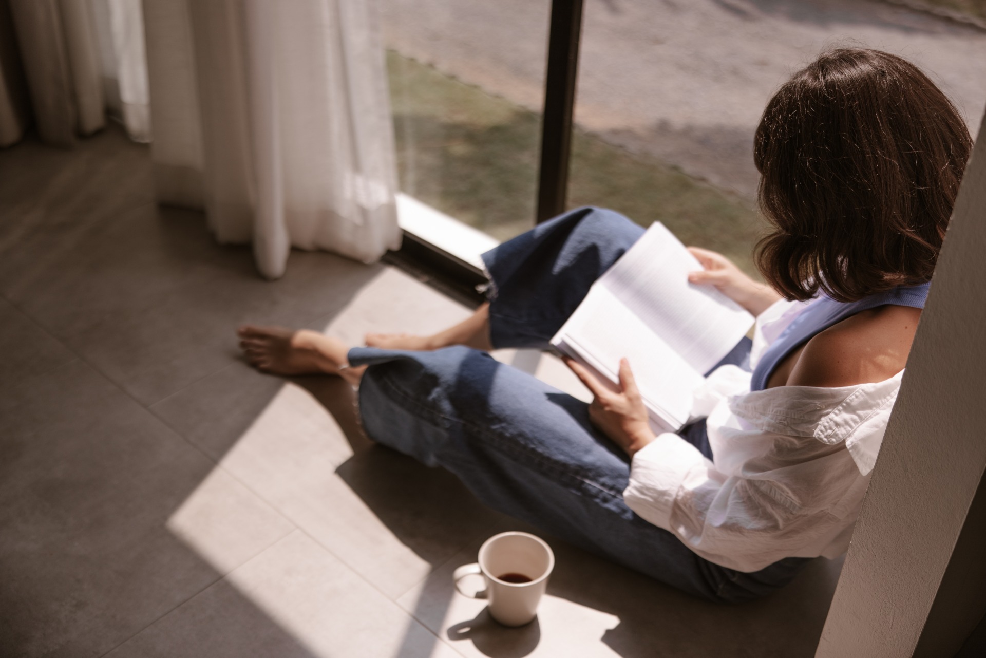 5 formas de conciliar vida e trabalho através do conceito Slow Living Vista superior de uma mulher sentada no chão perto de uma janela ensolarada, lendo um livro aberto com atenção. Ela está descalça, usando jeans largos e camisa branca. Uma caneca de café está no chão ao seu lado. Uma cena calma exemplificando o slow living.