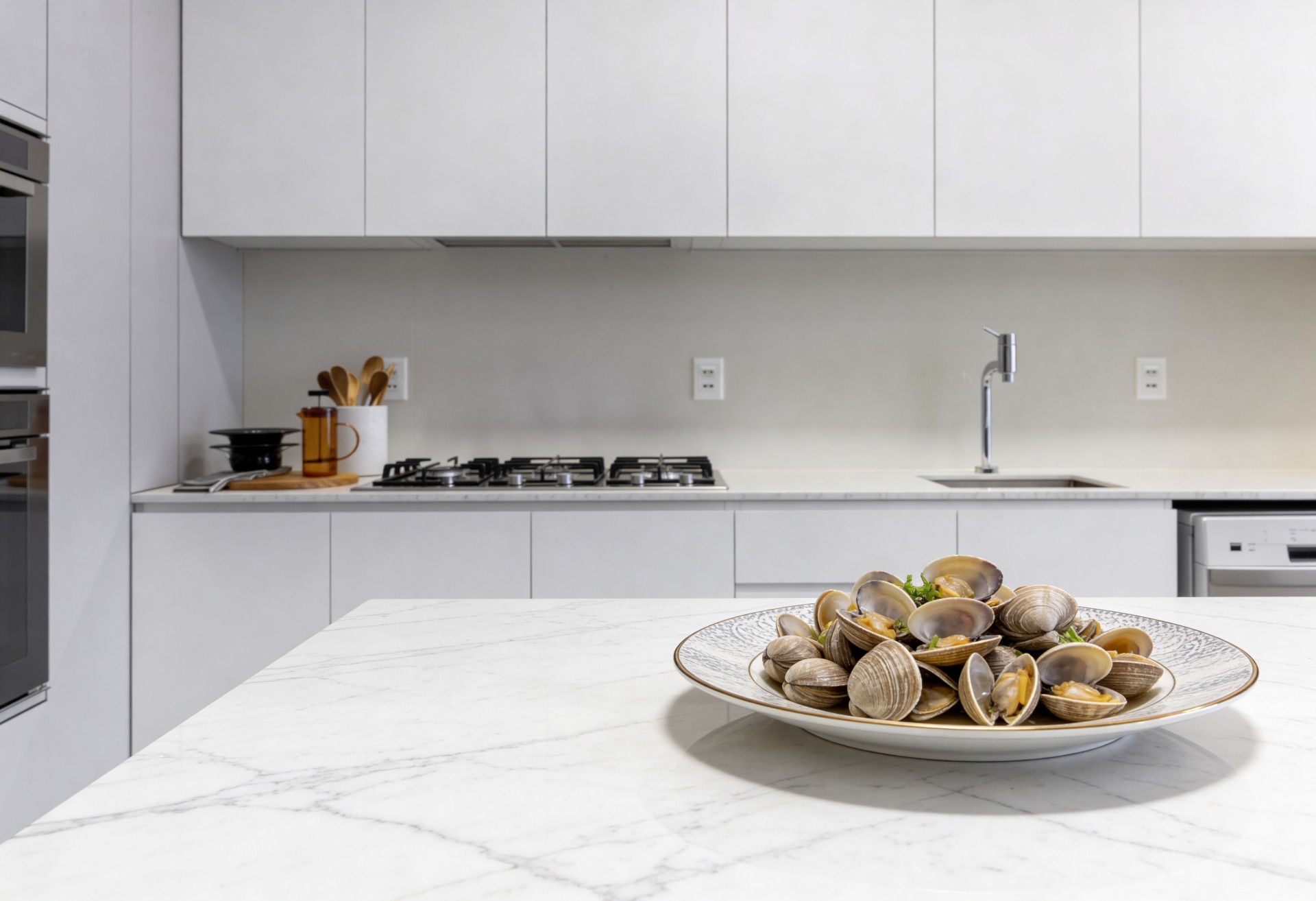 gastronomia-sensorial-berbigões-cozinha-minimalista Prato raso com berbigões cozidos em destaque sobre uma bancada de mármore branco. Ao fundo, uma cozinha moderna minimalista com armários brancos, fogão cooktop e utensílios de madeira.
