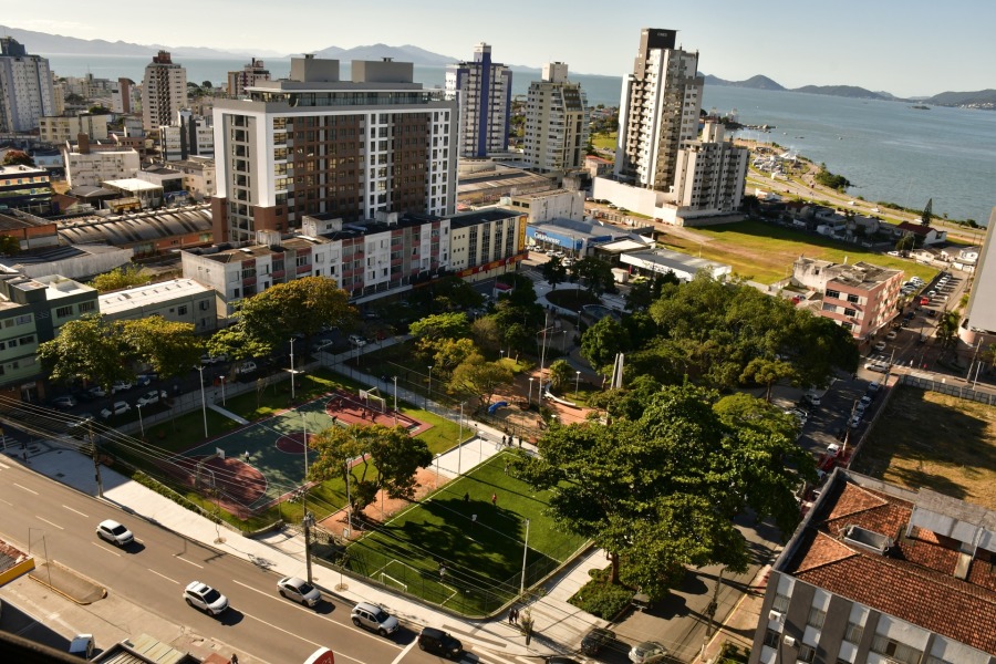 Fotografia aérea de uma praça urbana arborizada com uma quadra de basquete e um campo de futebol de grama sintética. Ao fundo, prédios residenciais e o mar sob um céu limpo.
