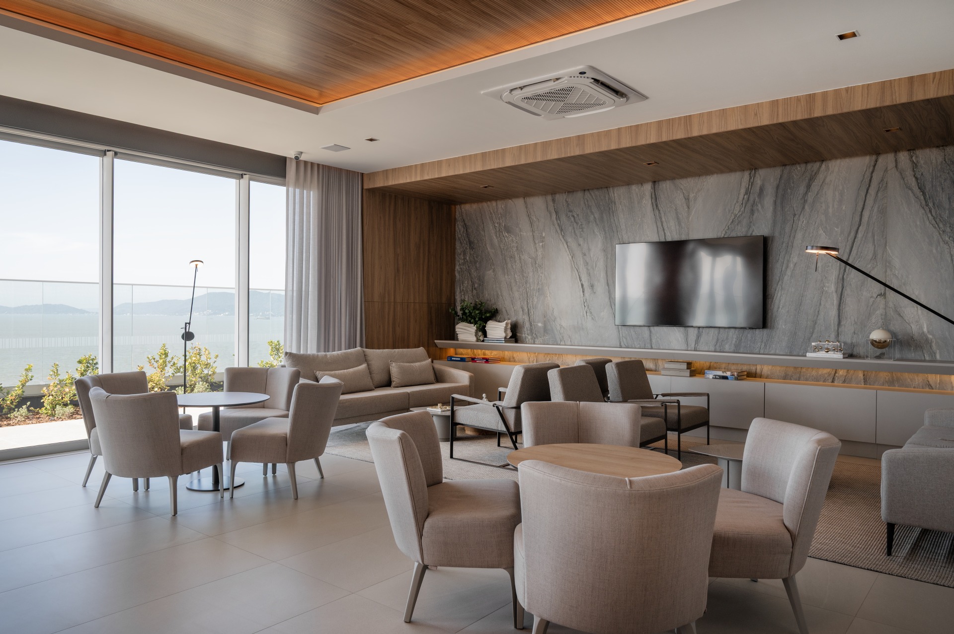 sala-lounge-moderna-vista-mar.jpg Fotografia em plano aberto de uma sala lounge ou área de convivência luxuosa e moderna. O ambiente possui diversas poltronas e cadeiras estofadas em tom bege claro, organizadas em pequenos grupos ao redor de mesas redondas de apoio. Ao fundo, há um grande painel de parede feito de mármore cinza com veios naturais, onde está fixada uma televisão de tela plana desligada. O teto e as laterais possuem acabamento em madeira clara com iluminação embutida. À esquerda, uma imensa parede de vidro do chão ao teto revela uma varanda com plantas e uma vista panorâmica para o mar e montanhas ao fundo, sob a luz do dia.